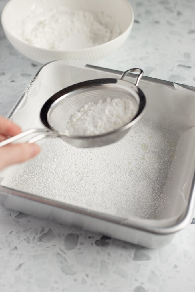 Hand sifting powdered sugar and cornstarch over a lined baking dish.