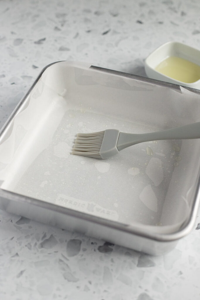 Pastry brush in a greased, lined baking tin, with a small bowl of oil in the background.
