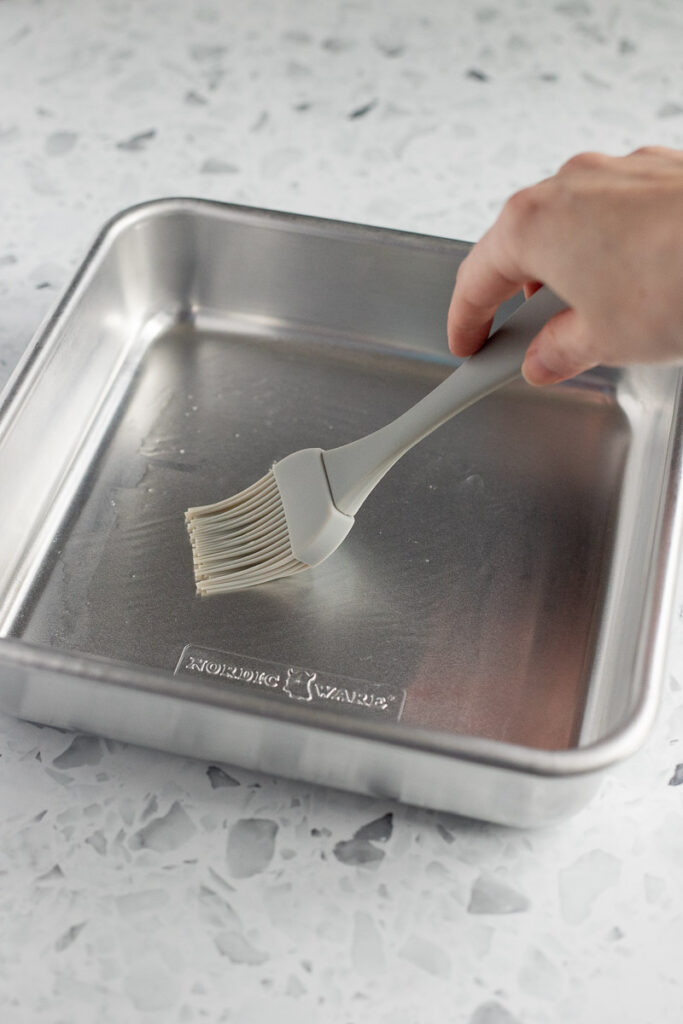 Hand greasing a metal baking dish with a pastry brush.