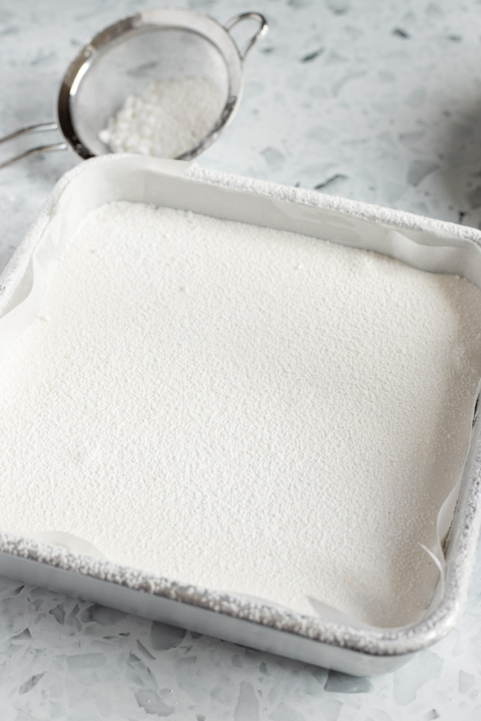 Homemade marshmallow in a baking dish, dusted with powdered sugar. A small sieve in the background.