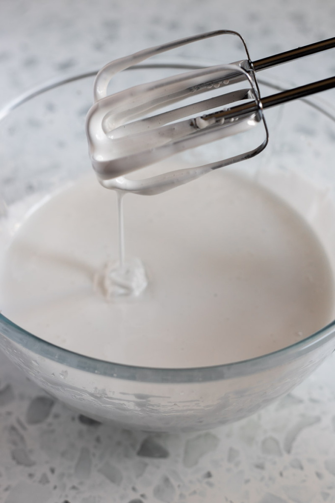 White, glossy marshmallow dripping from whisk into a clear mixing bowl.
