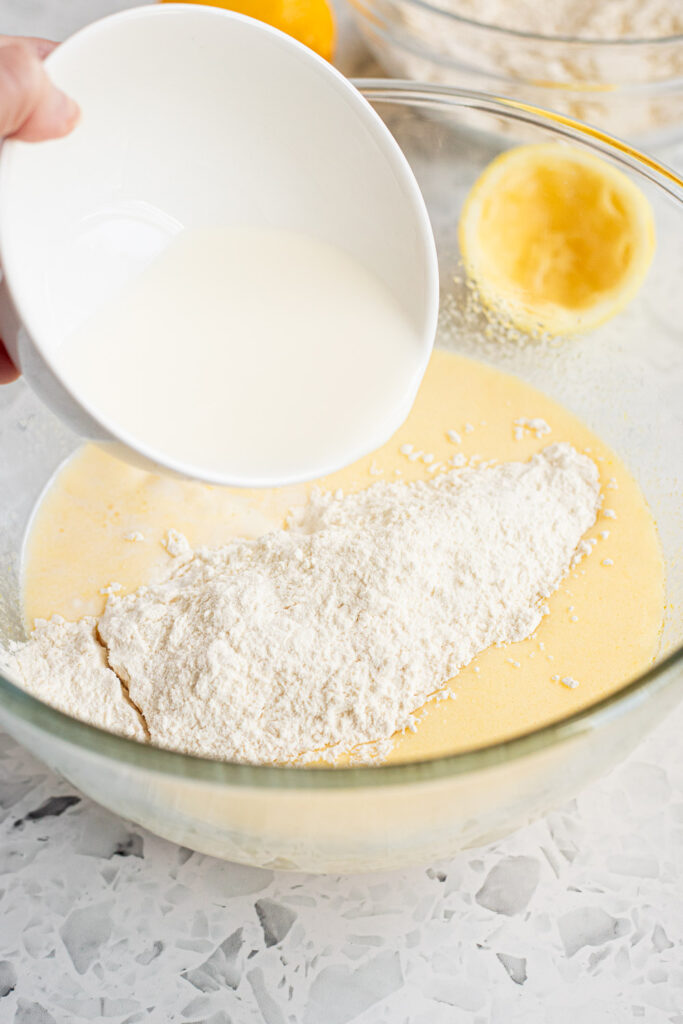 Milk being poured into lemon olive oil cake batter, with flour and other dry ingredients resting on the surface.