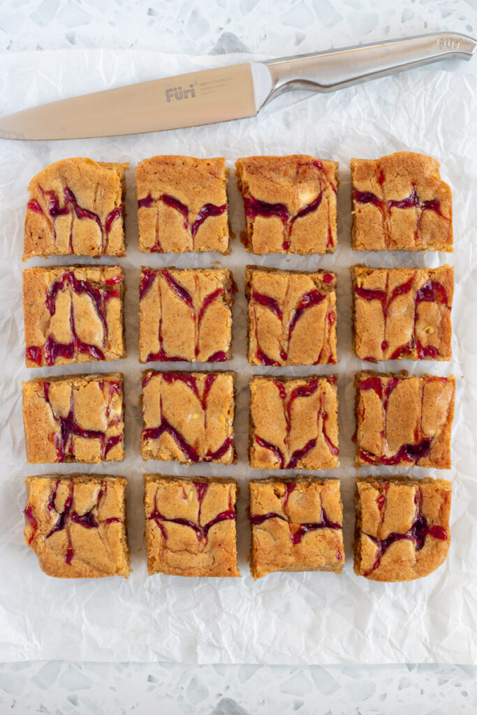 Jam blondies sliced into 16 squares on a piece of parchment paper. A long knife resting nearby.