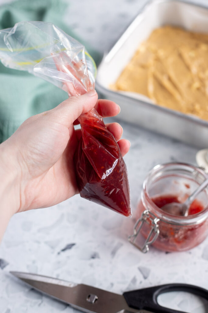 Hand holding a DIY piping bag filled with raspberry jam, with scissors and jam jar below.