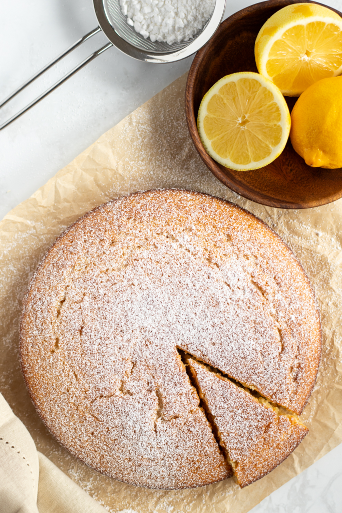 Italian lemon olive oil cake dusted with icing sugar, resting on crinkled brown parchment paper. A slice has been cut. Wooden bowl of lemons and a small sieve of icing sugar are in the background.