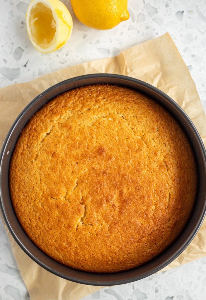 Baked lemon olive oil cake in a round metal cake tin lined with brown parchment paper. Golden brown with cracks on the surface.