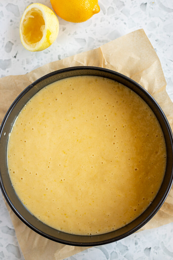 Round cake tin filled with lemon olive oil cake batter, bubbles visible on top, and brown parchment paper peeking out from underneath.