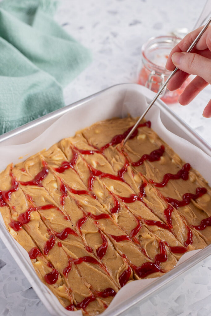 Hand dragging metal skewer vertically through lines of raspberry jam to form a chevron pattern.