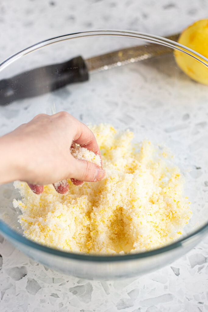 Hand rubbing lemon zest into granulated sugar with a microplane and zested lemon in the background. 