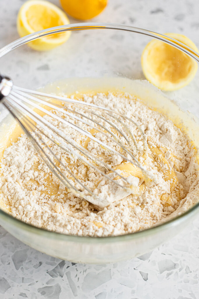 Mixing dry ingredients into wet lemon olive oil cake batter, with fresh halved lemons in the background.