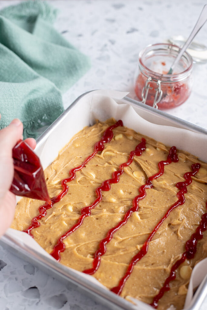 Hand piping horizontal lines of raspberry jam over white chocolate blondie batter.