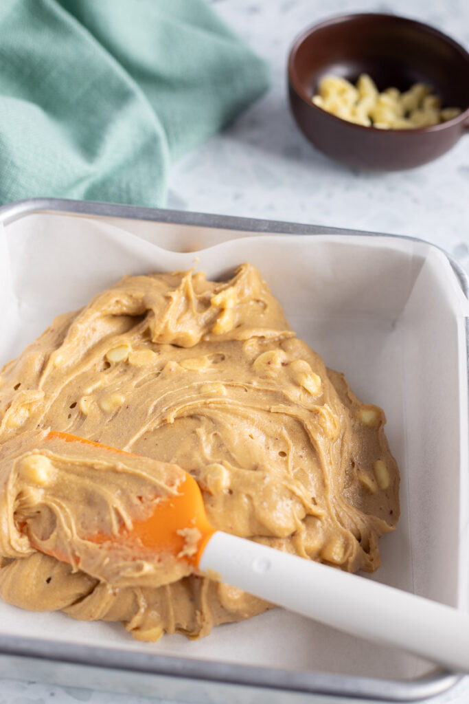 Spreading the blondie batter into a lined baking dish using a rubber spatula. A brown ramekin of chocolate chips nearby.