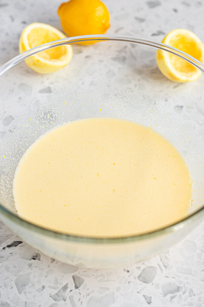 Wet cake ingredients, including lemon juice, vanilla, and eggs, whisked together in a mixing bowl. Halved lemons styled in the background.
