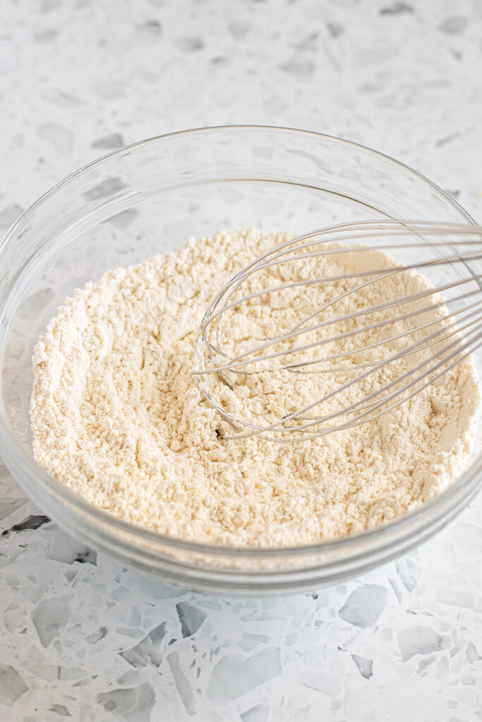 Flour, baking powder, and other dry ingredients mixed in a glass bowl with a metal whisk.