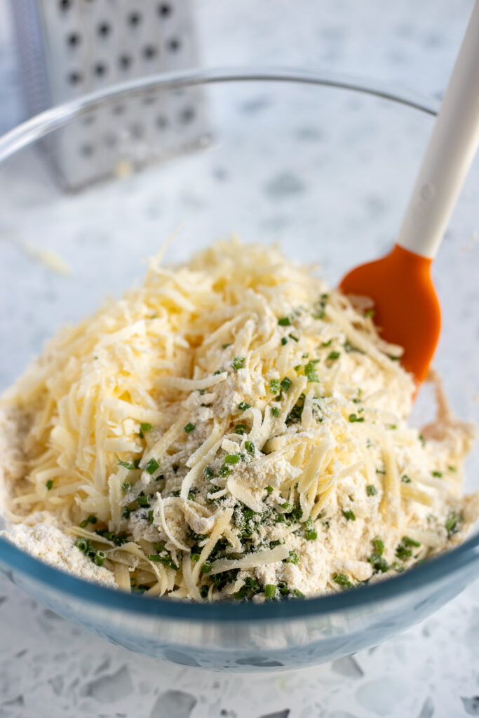 Shredded cheddar and chopped chives being stirred into flour mixture, with a metal cheese grater visible in the background.