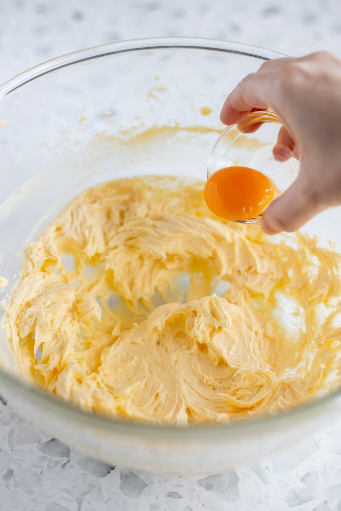 Hand adding egg yolk to a bowl of creamed butter and sugar.