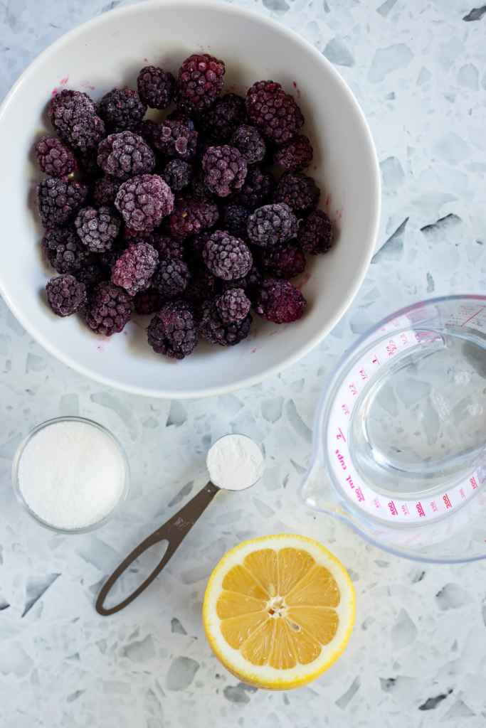 Overhead flat lay of simple blackberry sauce ingredients — frozen blackberries, lemon, sugar, cornstarch and water.