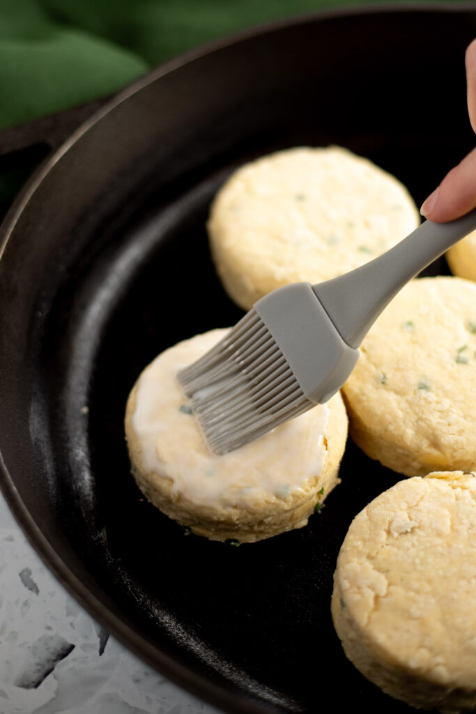Hand brushing buttermilk onto biscuits with a pastry brush, arranged in a cast iron skillet ready for baking.