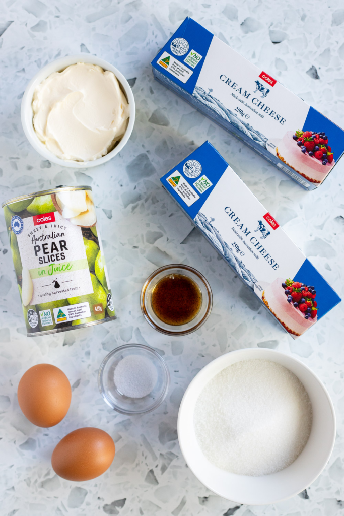 Overhead flat lay of ingredients for cheesecake filling, including cream cheese, canned pears, eggs, vanilla bean paste, sour cream and sugar.