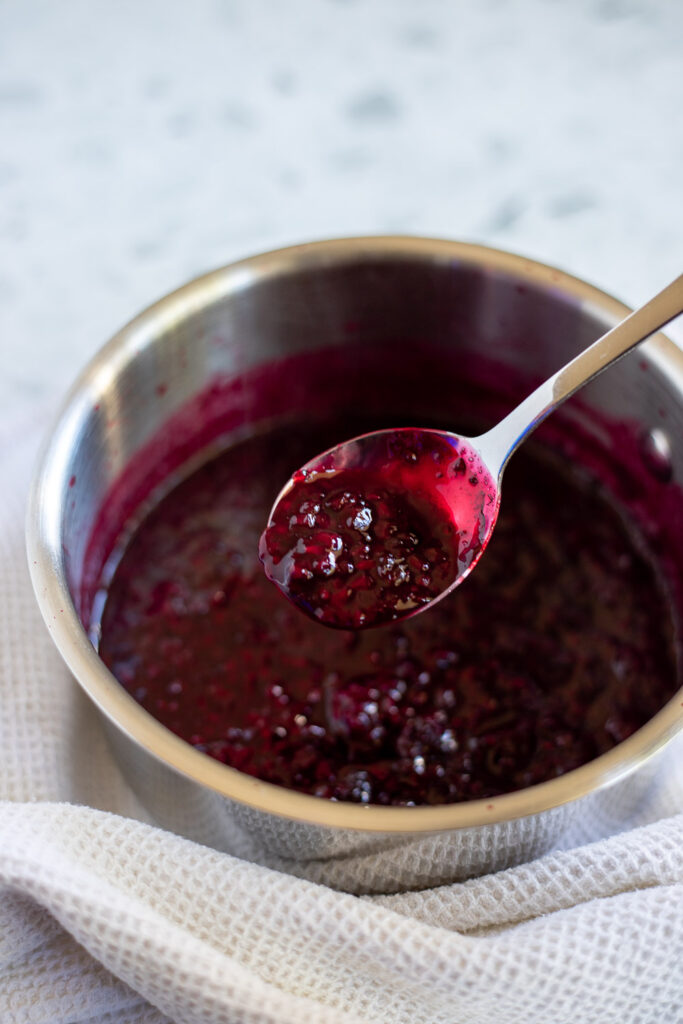 Saucepan of chunky blackberry sauce before straining, with a spoon dipped in the sauce and a white textured tea towel beside it