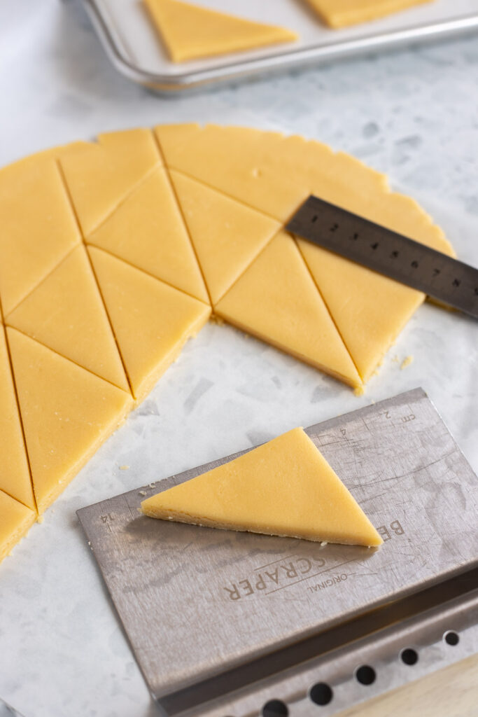 Shortbread dough cut into ‘bread slice’ triangles with a ruler as a guide; one triangle resting on a bench scraper, ready to transfer to baking tray.