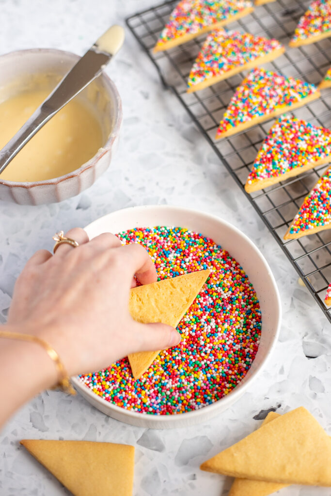 Hand pressing a shortbread cookie into a shallow bowl of colourful sprinkles, with a bowl of icing and a cooling rack of finished fairy shortbread in the background.