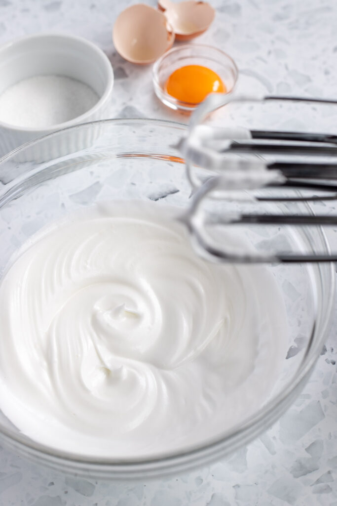 Egg whites whipped to stiff peaks in a mixing bowl, with beaters above, and a ramekin of sugar and an egg yolk in the background.