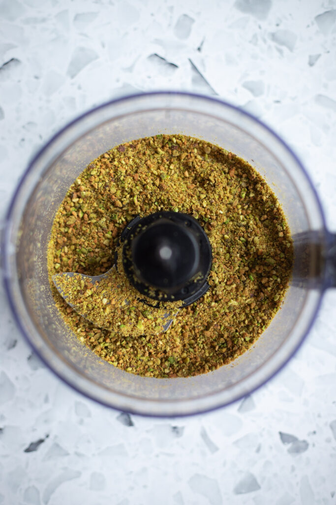 Top-down view of roasted pistachios ground into a coarse meal in a food processor for cupcake batter.