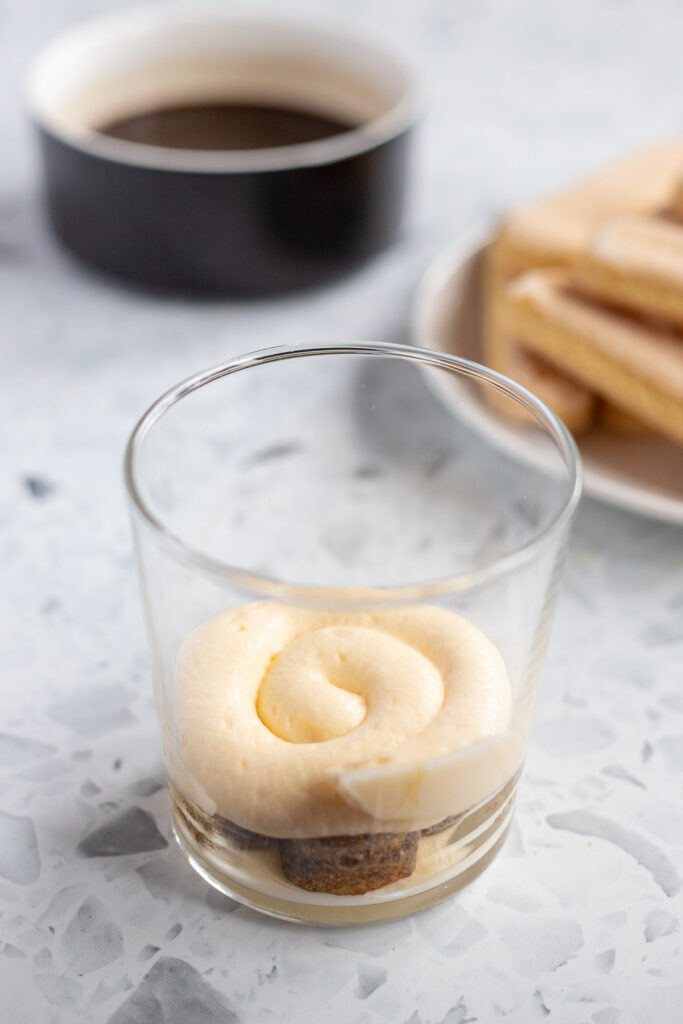 Close-up of tiramisu cup assembly, showing the second layer of smooth mascarpone cream over coffee-soaked ladyfingers.