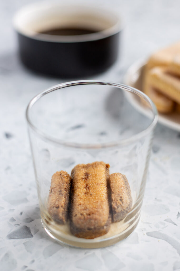 Close-up of assembling tiramisu in a cup, showing the first layer of coffee-soaked ladyfingers.