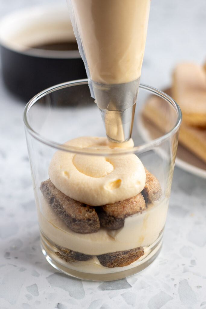 Close-up of piping the next layer of smooth mascarpone cream into a tiramisu cup over coffee-soaked ladyfingers.