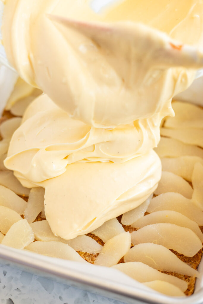 Close-up of creamy cheesecake filling being poured over neatly arranged pear slices in the baking tin.