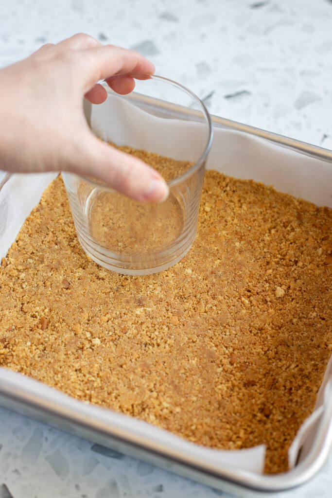 Using a glass to press the biscuit base evenly into a lined baking tin.