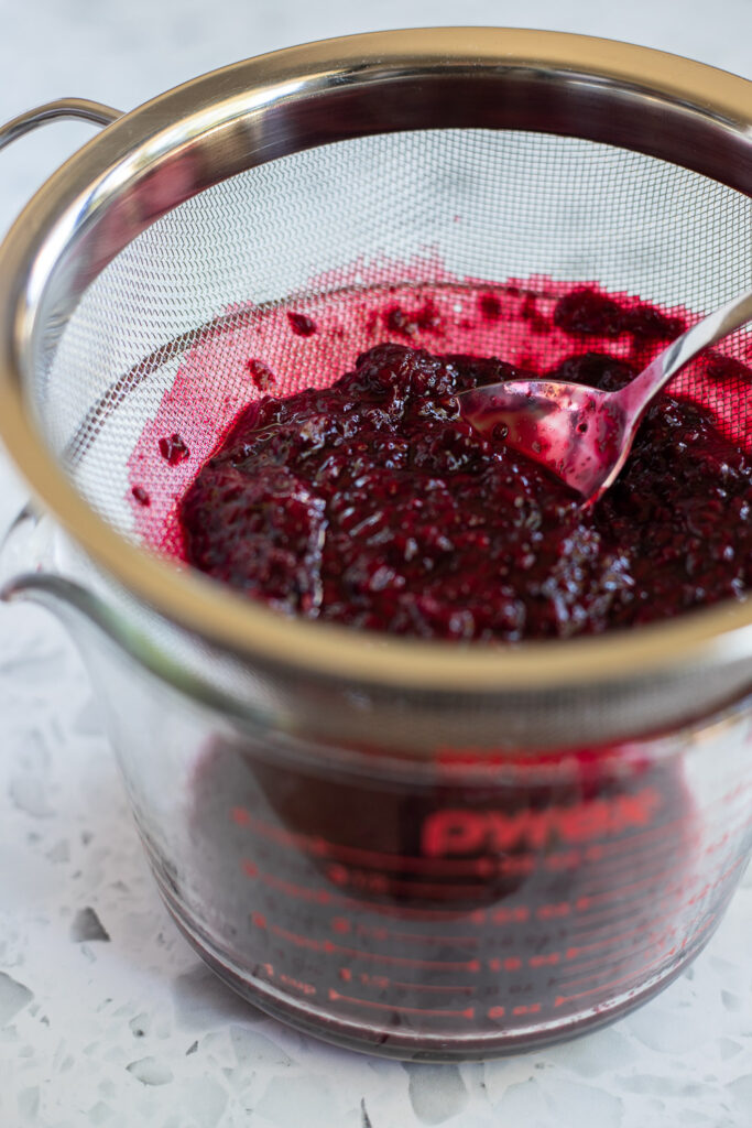 Close-up of straining homemade blackberry sauce through a fine mesh strainer into a Pyrex jug, with a spoon resting inside.