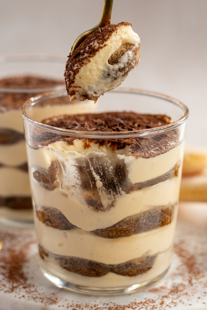 Close-up of a glass of tiramisu showing layers of coffee-soaked ladyfingers and creamy mascarpone, with a brass spoon holding a scoop of tiramisu above.