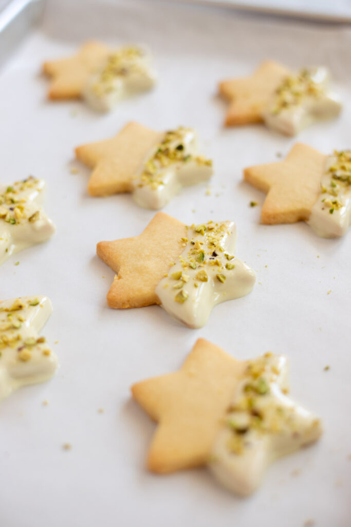 Lemon shortbread cookies half-dipped in white chocolate and sprinkled with pistachios, arranged on a baking tray ready to be refrigerated until set.