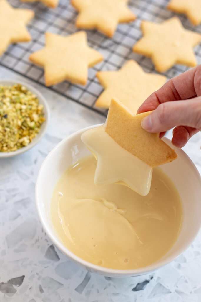 Hand half-dipping a lemon shortbread cookie into melted white chocolate, with a cooling rack of undecorated cookies in the background.