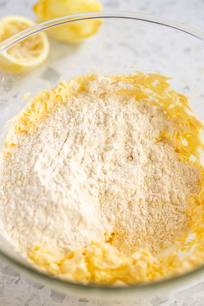 Flour on top of the shortbread butter mixture, with a squeezed lemon and a zested lemon in the background.
