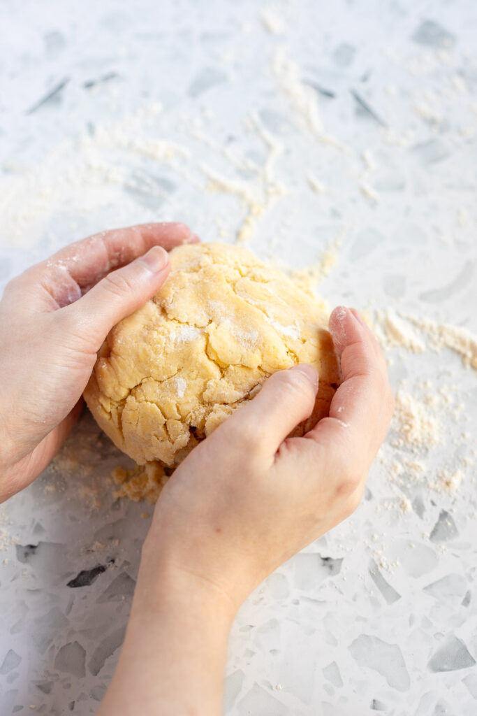 Hands forming crumbly dough into a ball on a floured work surface.