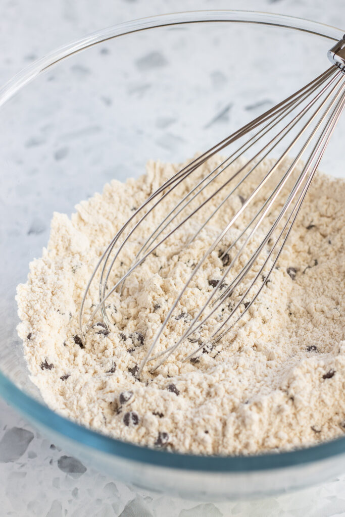 Bowl of combined dry ingredients, including chocolate chips, with a whisk resting inside.