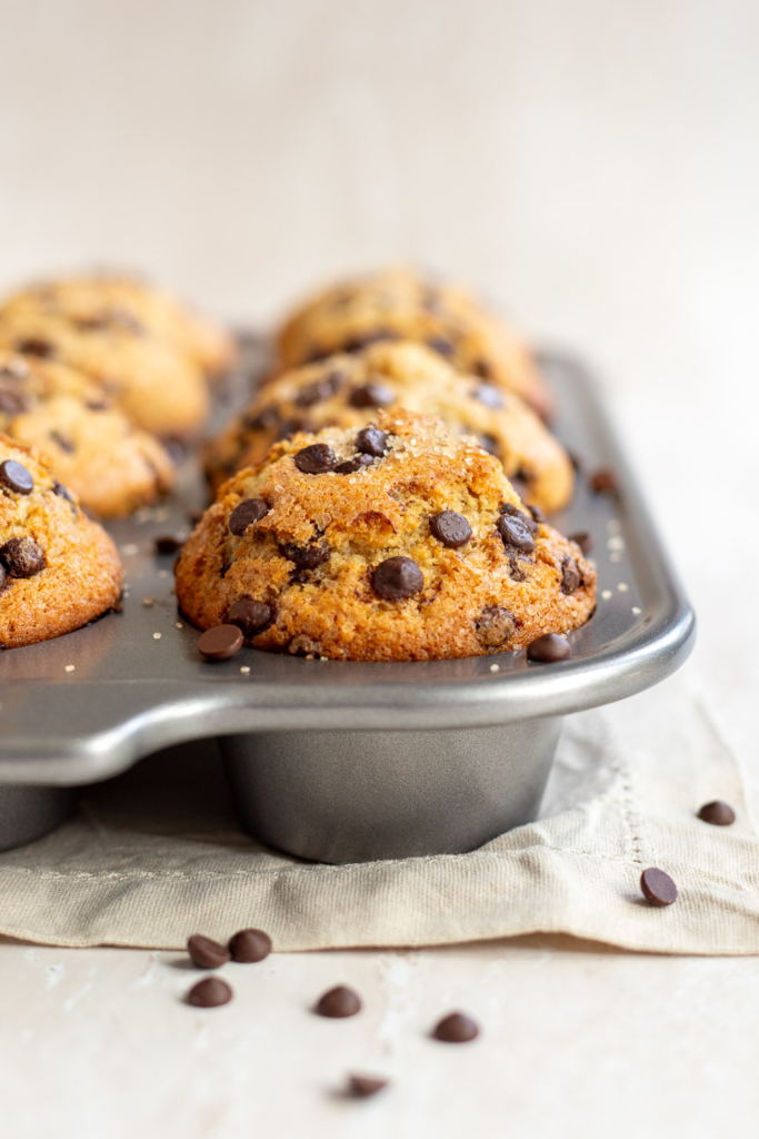 Straight-on view of bakery-style chocolate chip muffins in a tin, showing tall, golden domed tops.