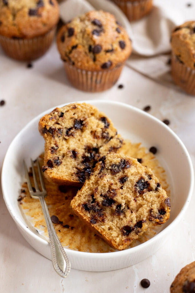 Bakery-style chocolate chip muffin cut in half in a shallow bowl, with more muffins and beige linens in the background.