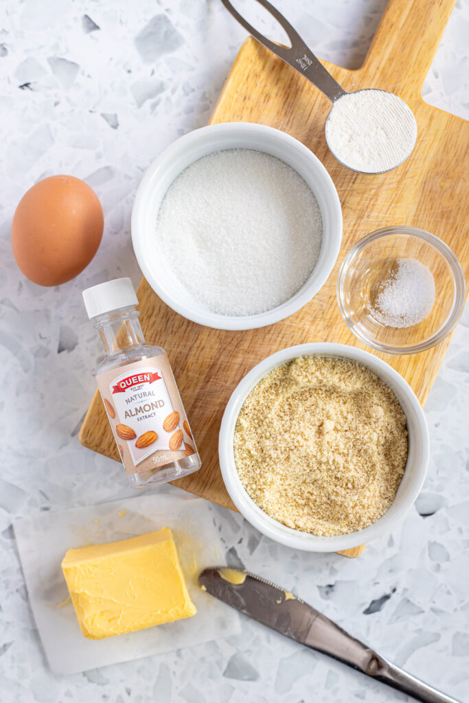 Flatlay of frangipane baking ingredients on a small wooden chopping board, including ground almonds, butter, sugar, and eggs.