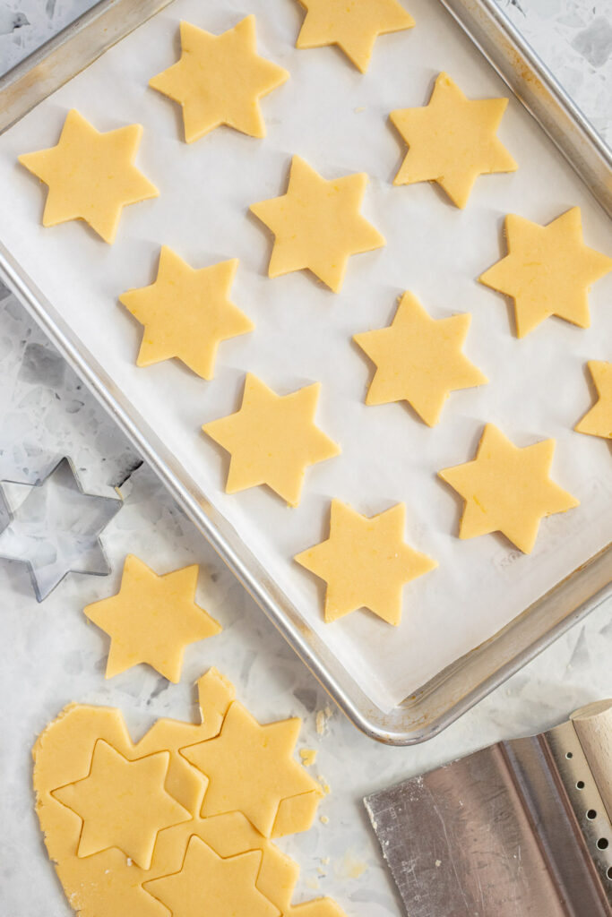 Overhead shot of star-shaped lemon shortbread cookies on a baking tray, cut from dough and transferred with a bench scraper.