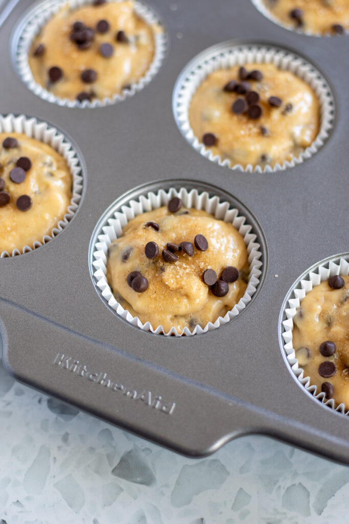 Chocolate chip muffin batter scooped into lined tin, nearly full, topped with coarse sugar and extra chocolate chips.