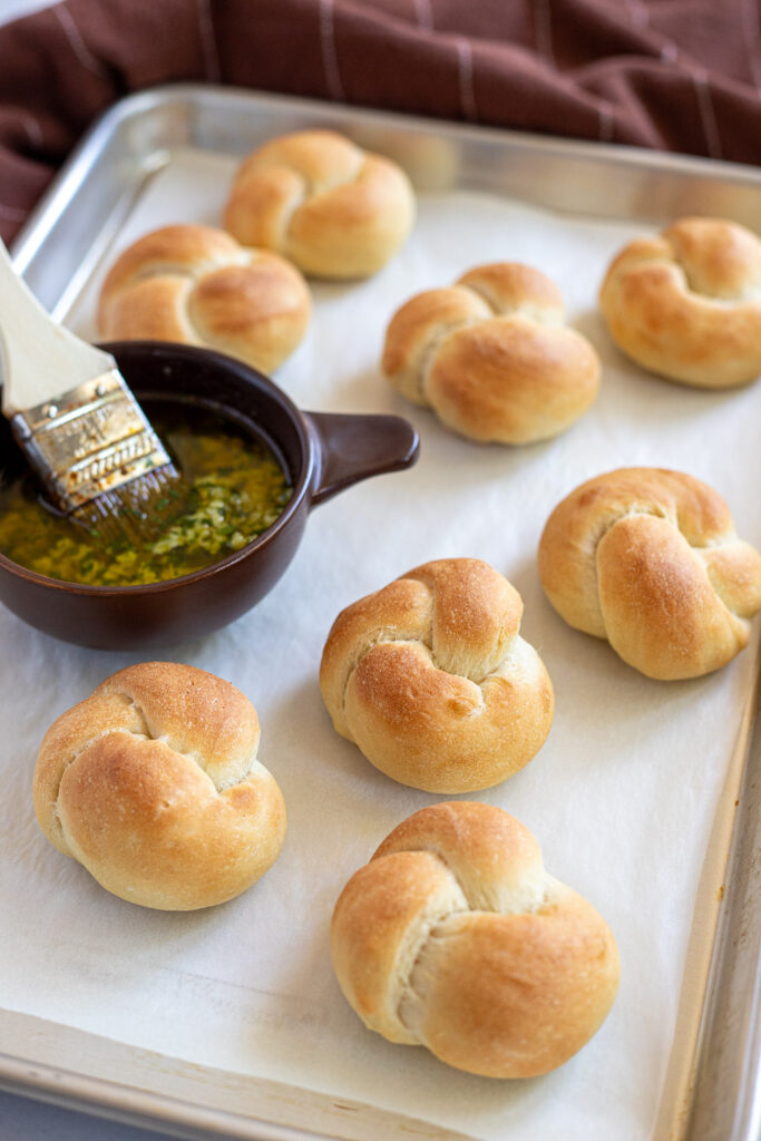 Freshly baked garlic knots on a tray with a ramekin of garlic butter and a pastry brush to the side.