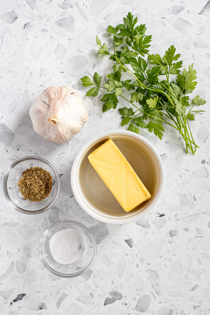 Ingredients for garlic butter on a countertop, including a garlic bulb, fresh parsley, and a stick of butter.