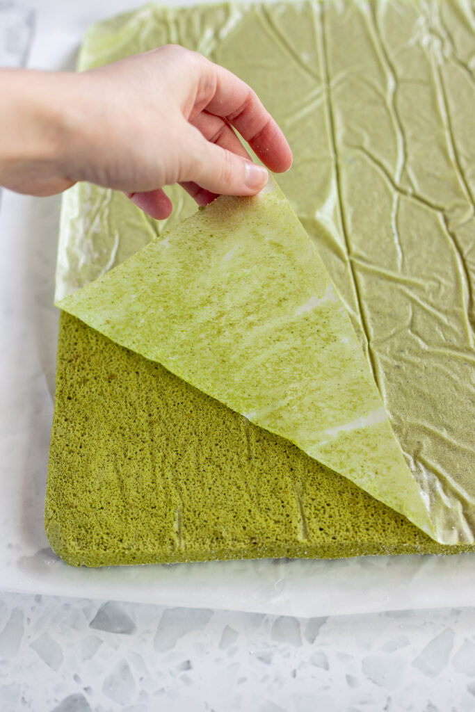 Peeling parchment paper off a matcha sponge after inverting it out of the baking tray.