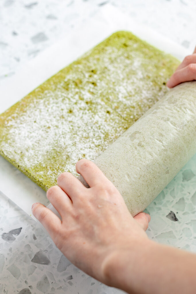 Hands pre-rolling matcha sponge in parchment paper to prevent cracking.