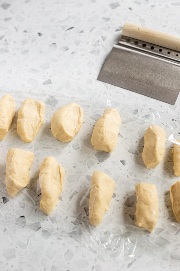Portioned garlic knot dough resting under cling film with a bench scraper in the background.